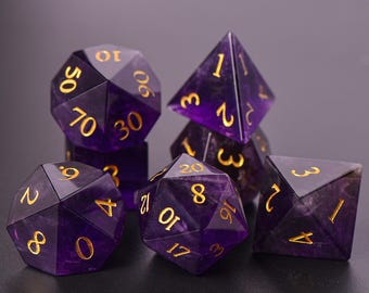 May include: Set of seven purple gemstone dice with gold numbering. The dice include a d4, d6, d8, d10, d12, d20, and a d100. The dice are arranged on a dark surface.