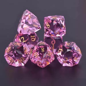 May include: A collection of seven pink, multi-faceted dice with gold numerals. The dice are displayed on a dark background, highlighting their geometric forms and reflective surfaces. Suitable for role-playing games.