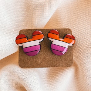 May include: A pair of earrings shaped like Mickey Mouse ears with a lesbian pride flag design. The earrings are red, orange, yellow, white, pink, and purple.