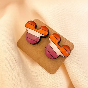 May include: A pair of wooden earrings shaped like Mickey Mouse ears. The earrings are painted with a pink, white, and orange striped pattern.