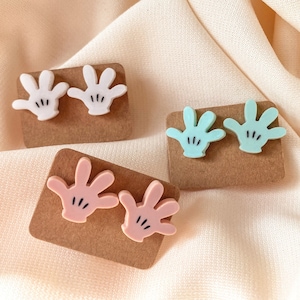 Puede incluir: Tres pares de peque&ntilde;os pendientes en forma de mano de colores. Los pendientes est&aacute;n sobre cartulina marr&oacute;n. Los pendientes son blancos, rosas y azul claro.