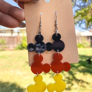 Puede incluir: Un par de pendientes colgantes con tres capas de orejas de Mickey Mouse. La capa superior es negra, la capa intermedia es roja y la capa inferior es amarilla.