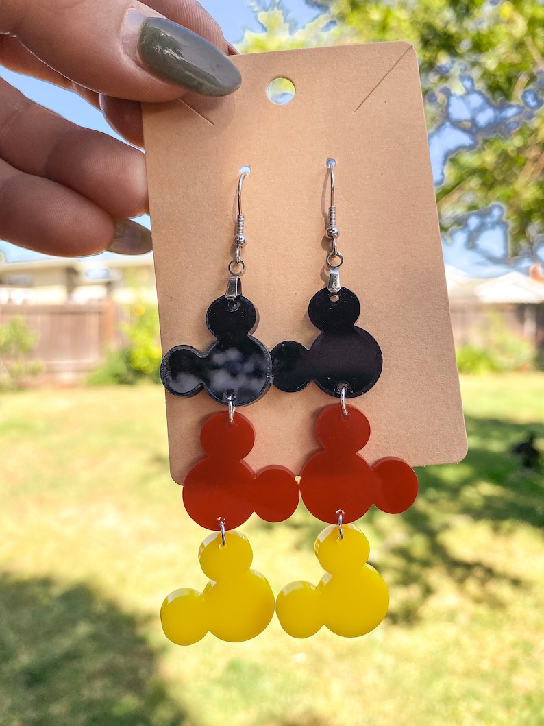 Puede incluir: Un par de pendientes colgantes con tres capas de orejas de Mickey Mouse. La capa superior es negra, la capa intermedia es roja y la capa inferior es amarilla.