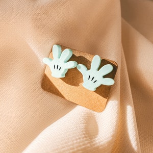 Puede incluir: Un par de pendientes de color azul claro en forma de mano con detalles negros. Los pendientes se muestran en una tarjeta marr&oacute;n.