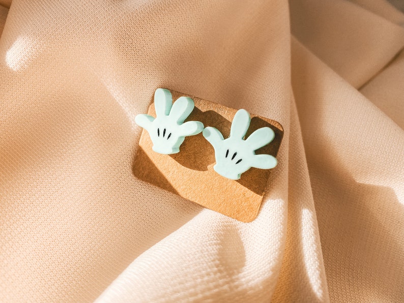 Puede incluir: Un par de pendientes de color azul claro en forma de mano con detalles negros. Los pendientes se muestran en una tarjeta marr&oacute;n.