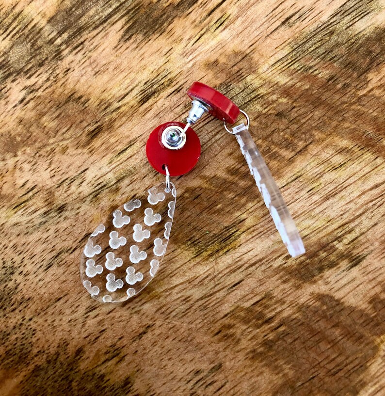 May include: Red and clear acrylic dangle earrings with a Mickey Mouse head pattern. The earrings have a red post and a clear acrylic dangle with a Mickey Mouse head pattern.