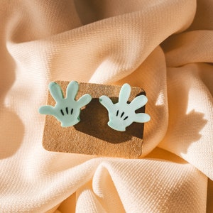 Puede incluir: Un par de pendientes de color azul claro en forma de mano con detalles negros. Los pendientes se muestran sobre una superficie marr&oacute;n.