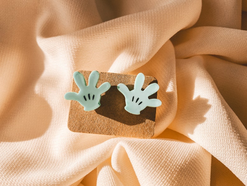 Puede incluir: Un par de pendientes de color azul claro en forma de mano con detalles negros. Los pendientes se muestran sobre una superficie marr&oacute;n.