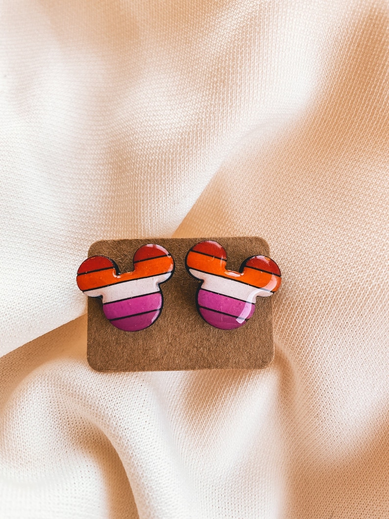May include: A pair of earrings shaped like Mickey Mouse ears with a lesbian pride flag design. The earrings are red, orange, yellow, white, pink, and purple.