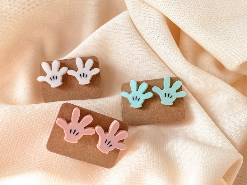 Puede incluir: Tres pares de peque&ntilde;os pendientes en forma de mano de colores. Los pendientes est&aacute;n sobre cartulina marr&oacute;n. Los pendientes son blancos, rosas y azul claro.