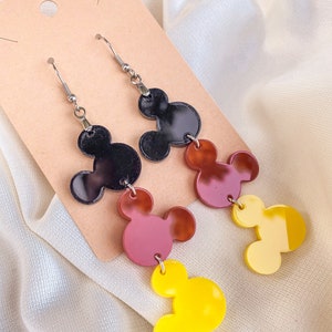 Puede incluir: Un par de pendientes colgantes con tres abalorios superpuestos en forma de cabeza de Mickey Mouse. El abalorio superior es negro, el abalorio del medio es marr&oacute;n y el abalorio inferior es amarillo.