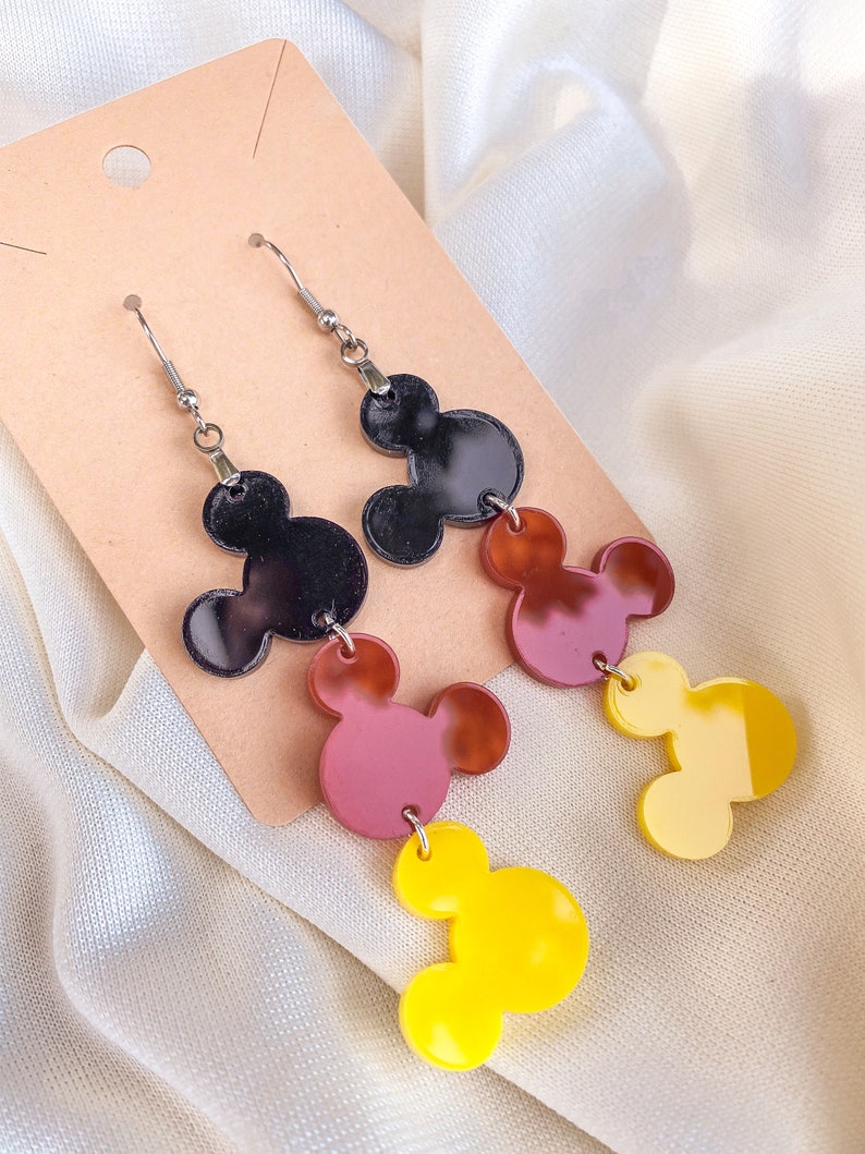 Puede incluir: Un par de pendientes colgantes con tres abalorios superpuestos en forma de cabeza de Mickey Mouse. El abalorio superior es negro, el abalorio del medio es marr&oacute;n y el abalorio inferior es amarillo.