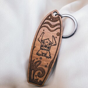 May include: Wooden keychain shaped like a surfboard with a cartoon character on it. The character is blue and has large eyes and a wide grin. The surfboard is brown and has a wave design.