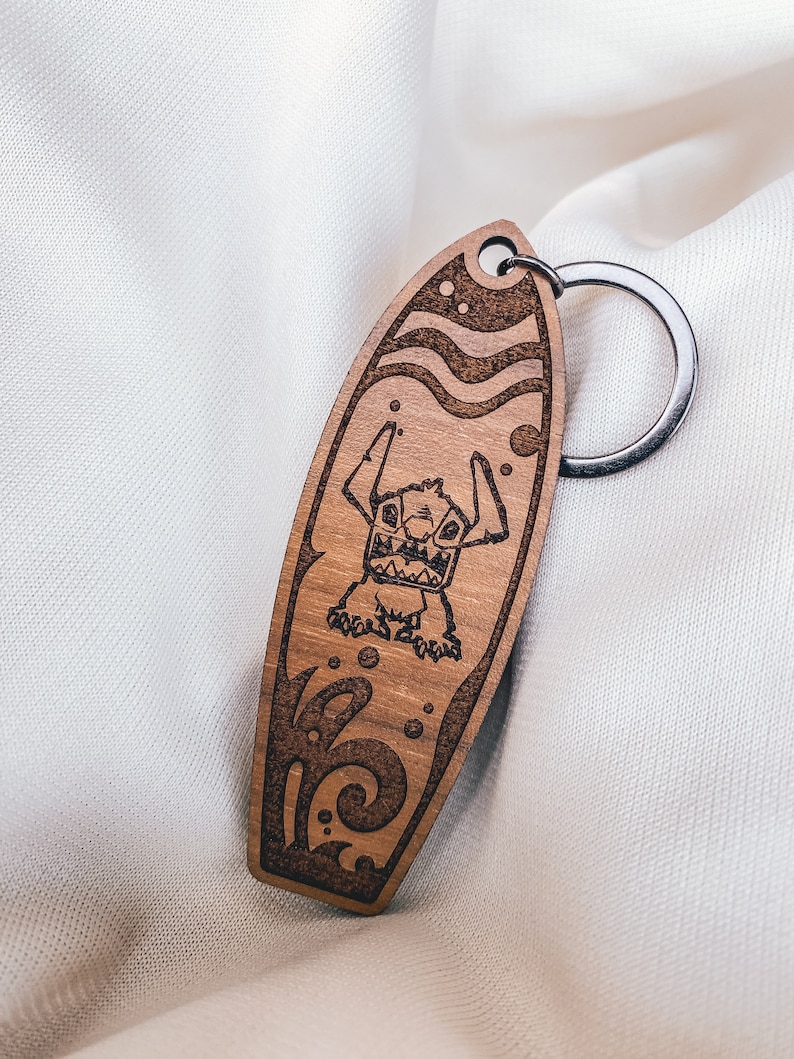 May include: Wooden keychain shaped like a surfboard with a cartoon character on it. The character is blue and has large eyes and a wide grin. The surfboard is brown and has a wave design.