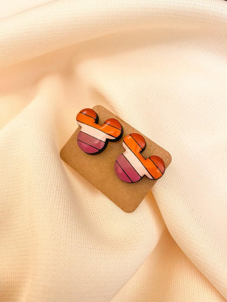May include: A pair of wooden earrings shaped like Mickey Mouse ears. The earrings are painted with a pink, white, and orange striped pattern.