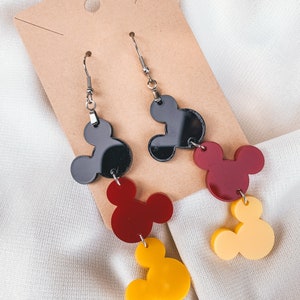 Puede incluir: Un par de pendientes colgantes con tres dijes de cabeza de Mickey Mouse superpuestos. El dije superior es negro, el dije del medio es rojo y el dije inferior es amarillo.