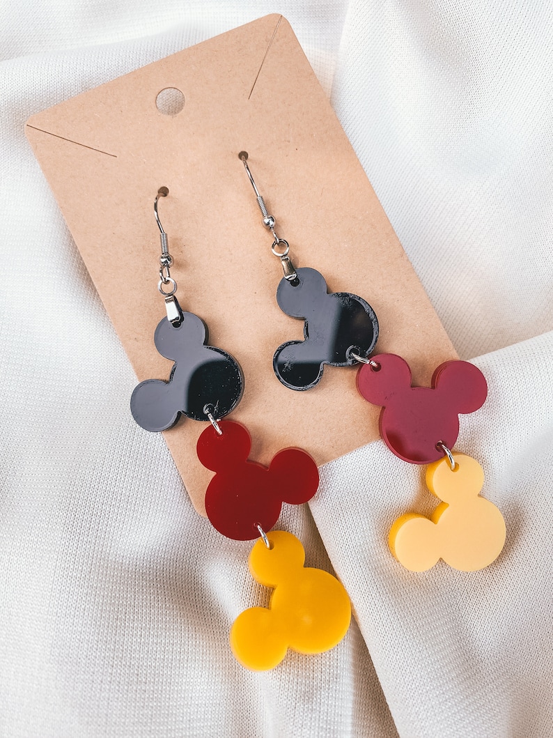 Puede incluir: Un par de pendientes colgantes con tres dijes de cabeza de Mickey Mouse superpuestos. El dije superior es negro, el dije del medio es rojo y el dije inferior es amarillo.