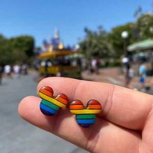 May include: A pair of rainbow-colored Mickey Mouse-shaped earrings. The earrings are made of wood and have a rainbow stripe pattern.