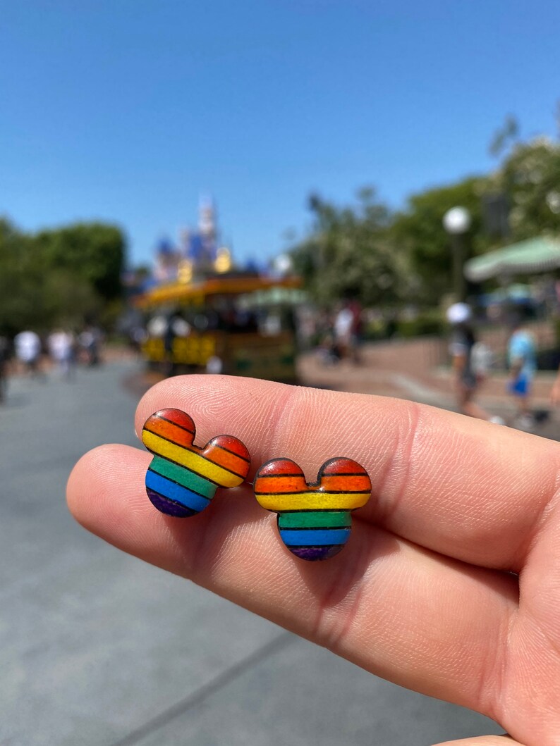 May include: A pair of rainbow-colored Mickey Mouse-shaped earrings. The earrings are made of wood and have a rainbow stripe pattern.