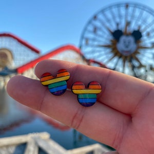 May include: A pair of rainbow-colored Mickey Mouse-shaped earrings. The earrings are made of wood and have a rainbow stripe pattern.