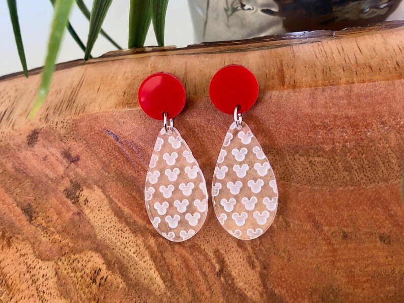 May include: A pair of red and clear dangle earrings. The clear teardrop-shaped earrings feature a white Mickey Mouse head pattern.