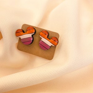May include: A pair of small, colorful, wooden earrings shaped like Mickey Mouse ears. The earrings have a striped design with orange, pink, and white stripes.