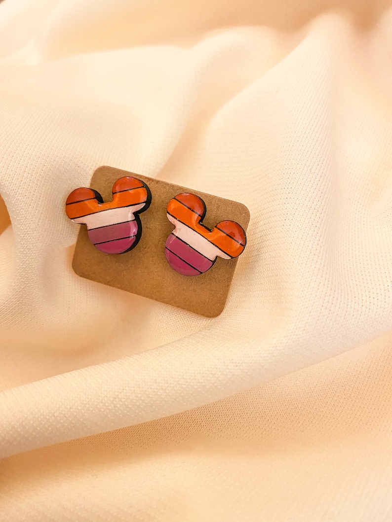 May include: A pair of small, colorful, wooden earrings shaped like Mickey Mouse ears. The earrings have a striped design with orange, pink, and white stripes.