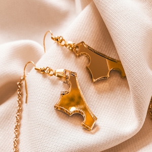 May include: A pair of gold earrings with a unique geometric design. The earrings are made of metal and have a mirror-like finish. They are hanging on a white fabric background.