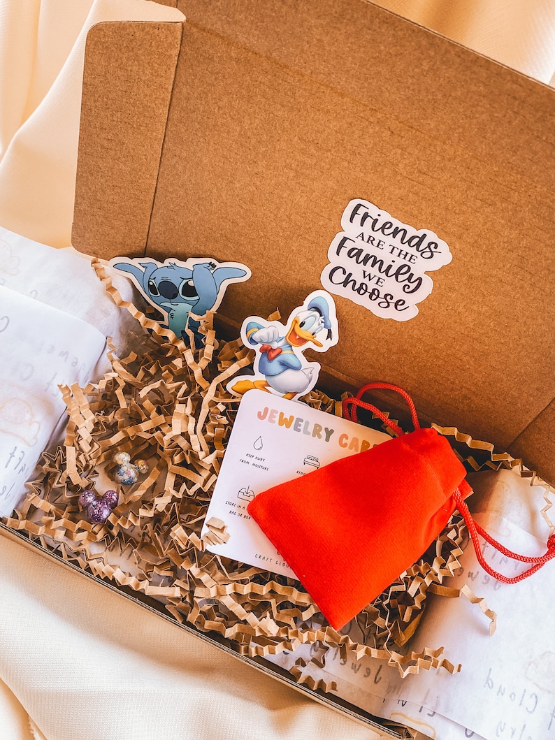 May include: A brown cardboard box filled with brown packing paper. The box contains a red pouch, two small colorful earrings, a card with the text "Jewelry Care" and three stickers. One sticker features Stitch from Lilo & Stitch, another features Donald Duck, and the third sticker says "Friends are the family we choose."