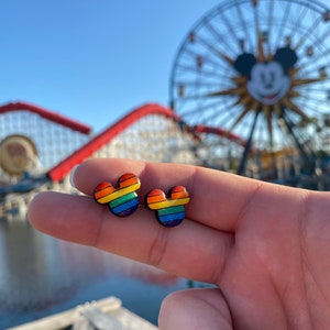 May include: A pair of rainbow Mickey Mouse-shaped earrings. The earrings are made of a colorful material and have a black outline.