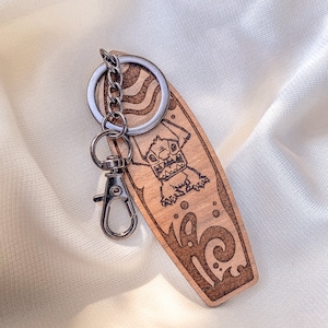May include: A wooden surfboard keychain with a cartoon monster surfing on a wave. The keychain is brown with a silver metal ring and clasp.
