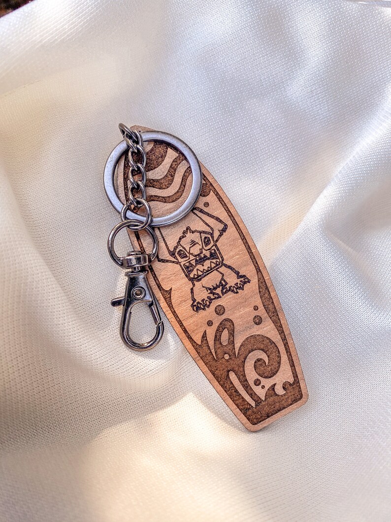 May include: A wooden surfboard keychain with a cartoon monster surfing on a wave. The keychain is brown with a silver metal ring and clasp.