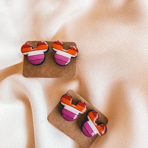 May include: A pair of lesbian pride flag Mickey Mouse earrings on a brown card. The earrings are red, orange, yellow, white, pink, and purple.