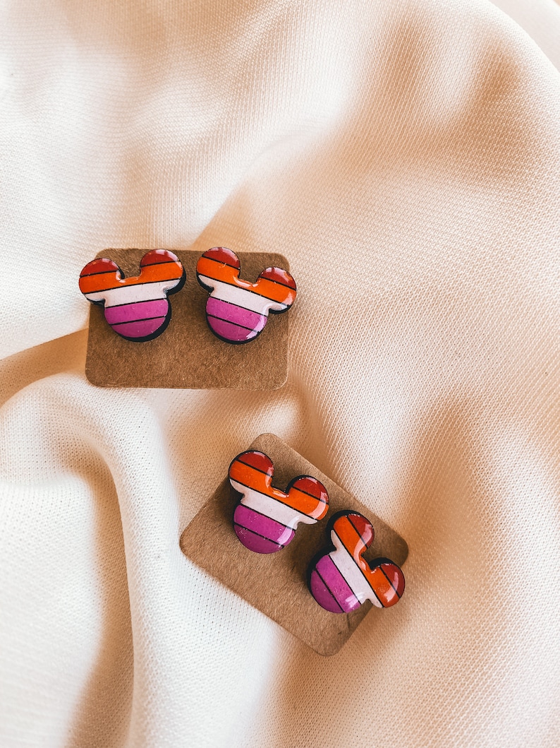 May include: A pair of lesbian pride flag Mickey Mouse earrings on a brown card. The earrings are red, orange, yellow, white, pink, and purple.