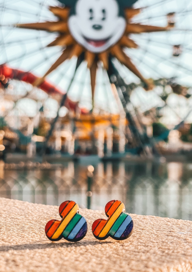May include: A pair of rainbow Mickey Mouse-shaped earrings. The earrings are made of a colorful, striped material and are in focus against a blurred background of a Ferris wheel.