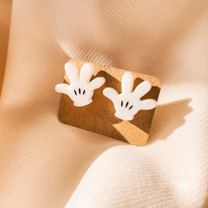 Puede incluir: Un par de pendientes blancos con forma de mano de dibujos animados en una tarjeta de presentaci&oacute;n marr&oacute;n. Las manos tienen detalles negros para los dedos y el pulgar.