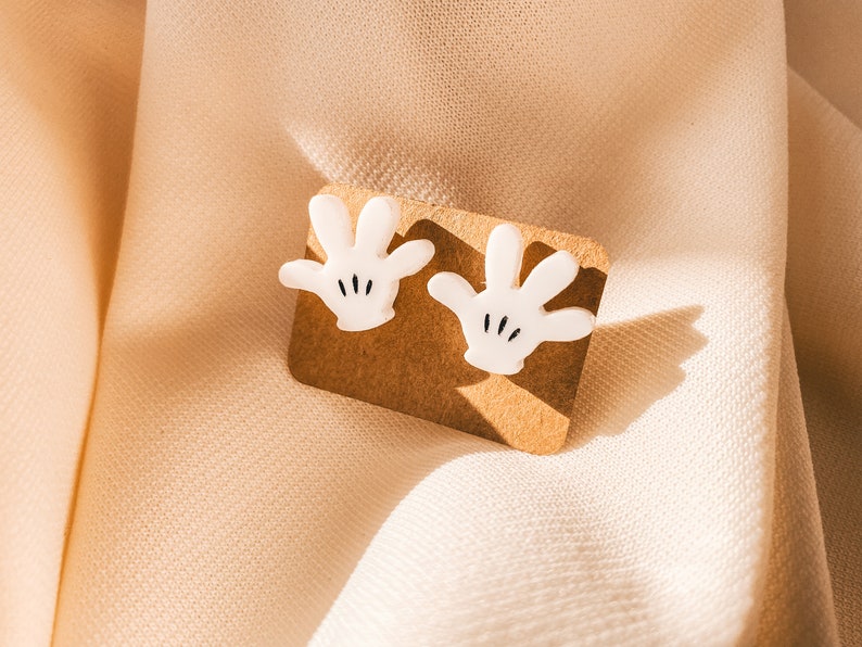 Puede incluir: Un par de pendientes blancos con forma de mano de dibujos animados en una tarjeta de presentaci&oacute;n marr&oacute;n. Las manos tienen detalles negros para los dedos y el pulgar.