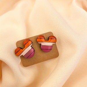 May include: A pair of small, colorful striped Mickey Mouse-shaped earrings. The earrings are made of a pink, orange, and white striped material.