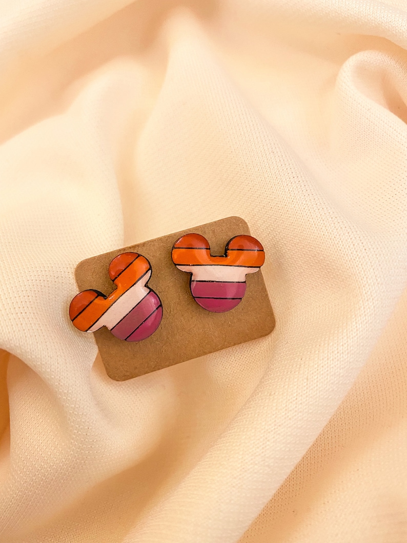 May include: A pair of small, colorful striped Mickey Mouse-shaped earrings. The earrings are made of a pink, orange, and white striped material.