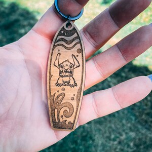 May include: Wooden surfboard keychain with a cartoon Stitch character from Lilo & Stitch. The keychain features a brown wooden surfboard with a blue and white wave design and a black metal ring.
