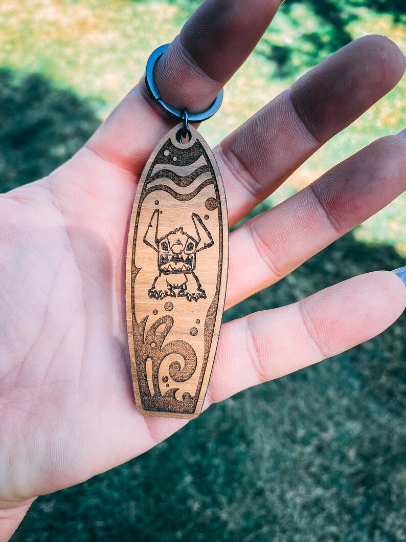 May include: Wooden surfboard keychain with a cartoon Stitch character from Lilo & Stitch. The keychain features a brown wooden surfboard with a blue and white wave design and a black metal ring.