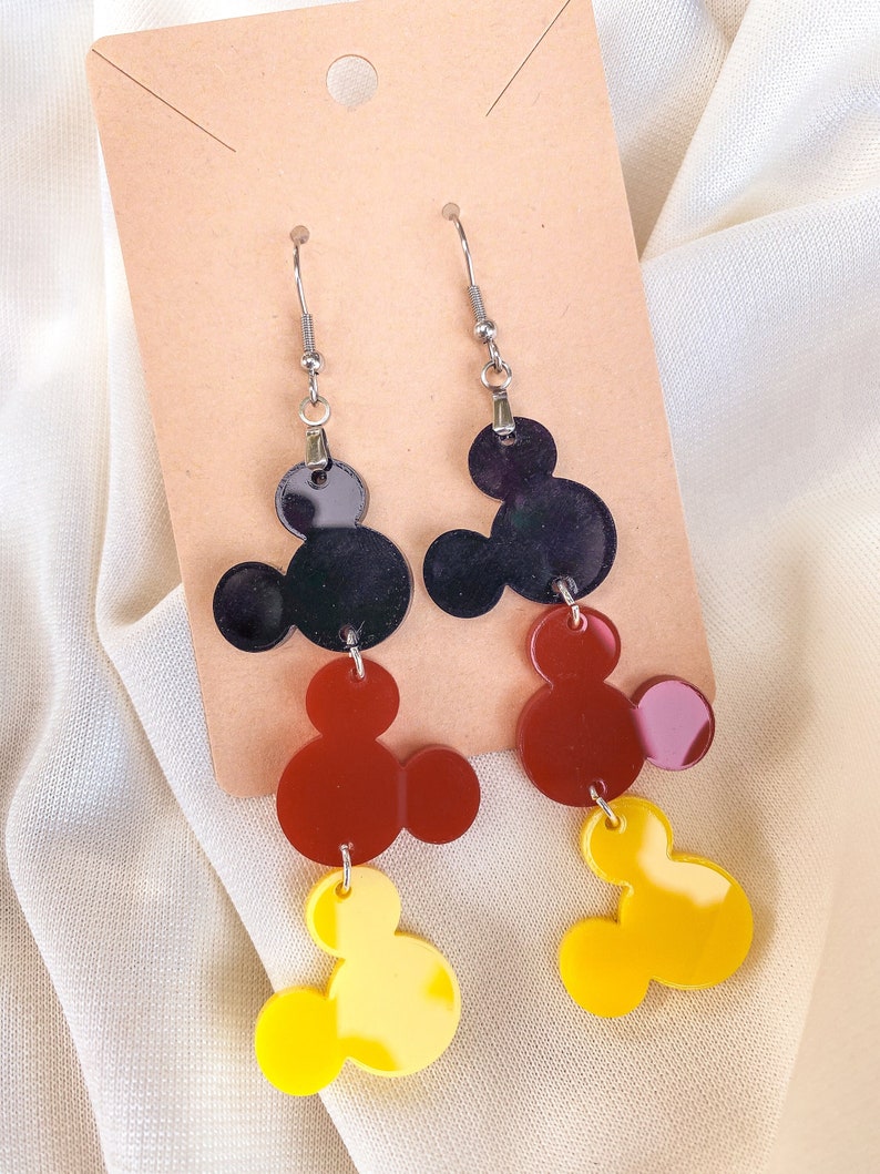 Puede incluir: Un par de pendientes colgantes con tres dijes de cabeza de Mickey Mouse superpuestos. El dije superior es negro, el dije del medio es rojo y el dije inferior es amarillo.