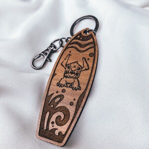 May include: A wooden surfboard keychain with a cartoon Stitch character from Lilo & Stitch. The keychain is brown with a black outline and features a wave design.