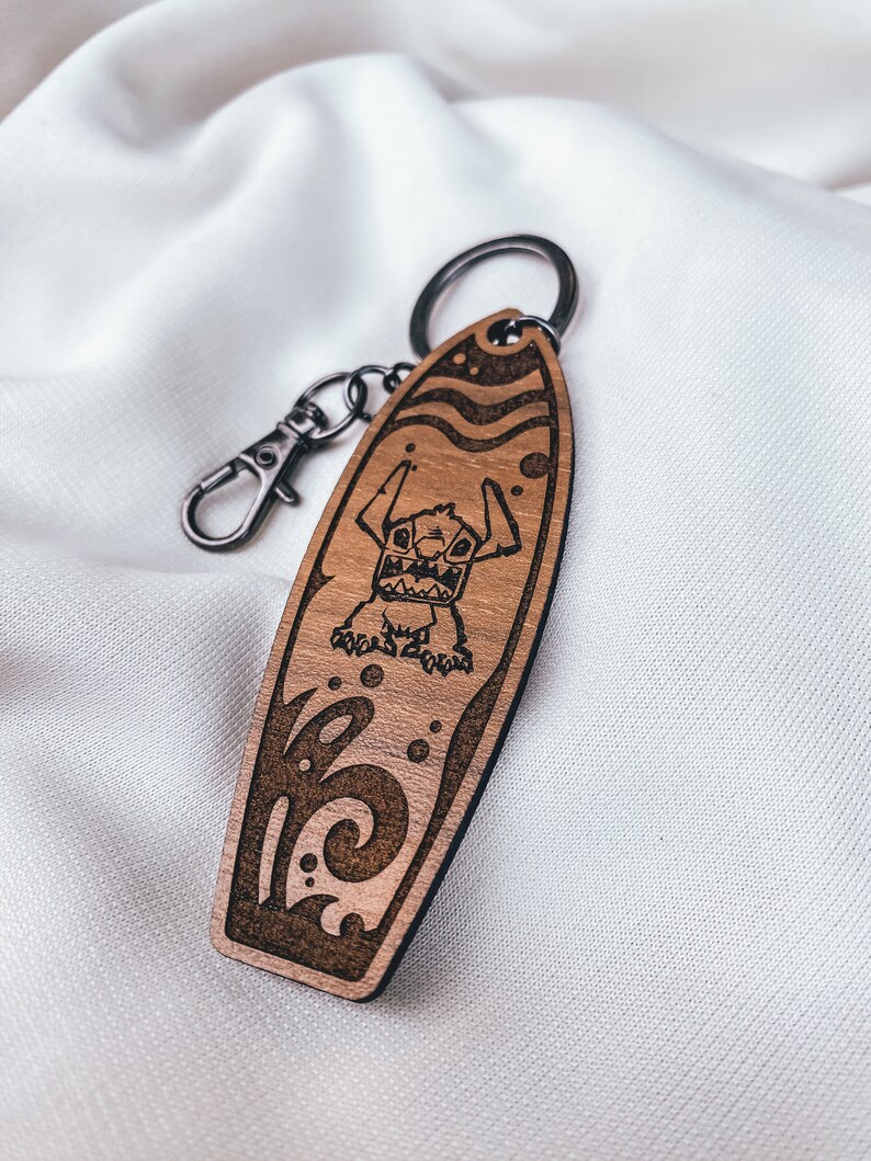 May include: A wooden surfboard keychain with a cartoon Stitch character from Lilo & Stitch. The keychain is brown with a black outline and features a wave design.