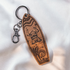 May include: Wooden surfboard keychain with a cartoon character on it. The character is a blue creature with large eyes and a toothy grin. The keychain is brown and has a silver ring.