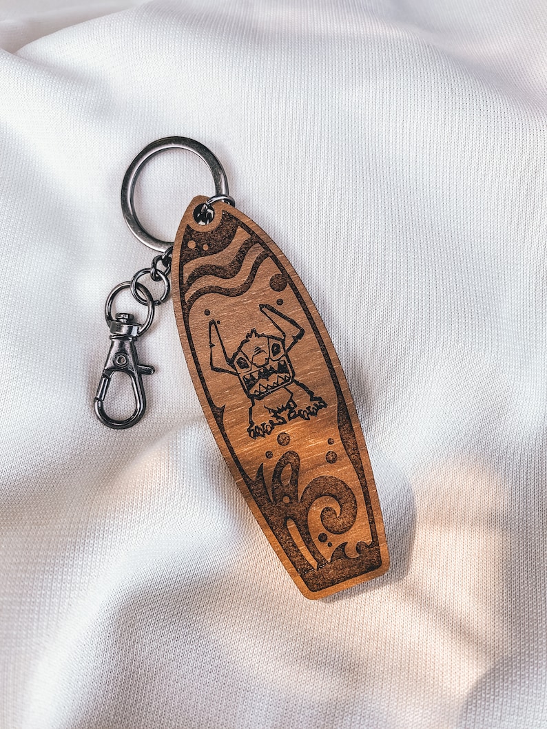 May include: Wooden surfboard keychain with a cartoon character on it. The character is a blue creature with large eyes and a toothy grin. The keychain is brown and has a silver ring.