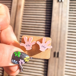 Puede incluir: Un par de pendientes de bot&oacute;n de mano rosa con detalles negros. Los pendientes se muestran en una tarjeta marr&oacute;n.