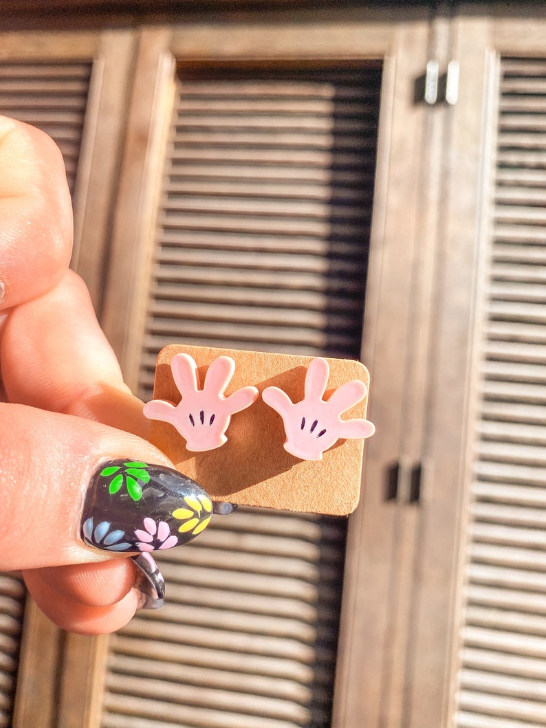 Puede incluir: Un par de pendientes de bot&oacute;n de mano rosa con detalles negros. Los pendientes se muestran en una tarjeta marr&oacute;n.