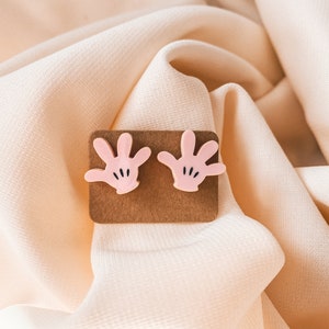 Puede incluir: Un par de pendientes de mano rosa de estilo de dibujos animados con detalles negros. Los pendientes se muestran en una tarjeta marr&oacute;n.