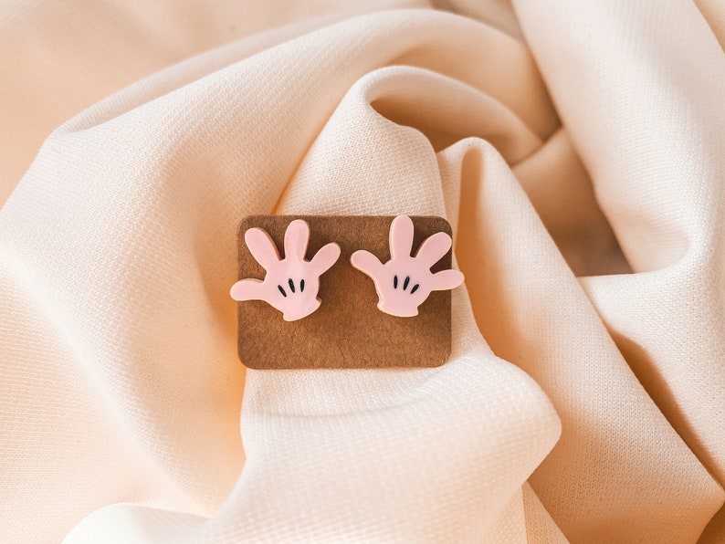 Puede incluir: Un par de pendientes de mano rosa de estilo de dibujos animados con detalles negros. Los pendientes se muestran en una tarjeta marr&oacute;n.